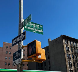 New York City street signs on Amsterdam Ave and at 167th St in Washington Heights. Behind the street signs, residential buildings rise from the ground and into the cobalt blue sky.