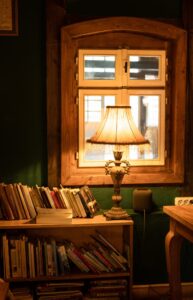 a bookshelf next to a window and warmly lit lamp