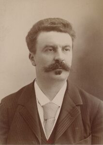 a sepia-toned portrait photograph of a white man wearing a suit and tie and sporting a large mustache and small goatee