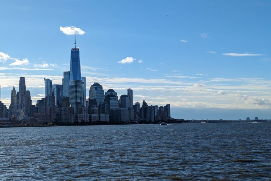 "Photograph of Lower Manhattan showcasing the One World Trade Center, as viewed from a ferry on the Hudson River. The skyscraper dominates the skyline, standing tall amidst surrounding buildings. The water of the Hudson River is visible in the foreground.The image captures the urban landscape and might include elements like the blue sky or other buildings, offering a perspective of the city's architecture and its relationship with the natural environment of the river."