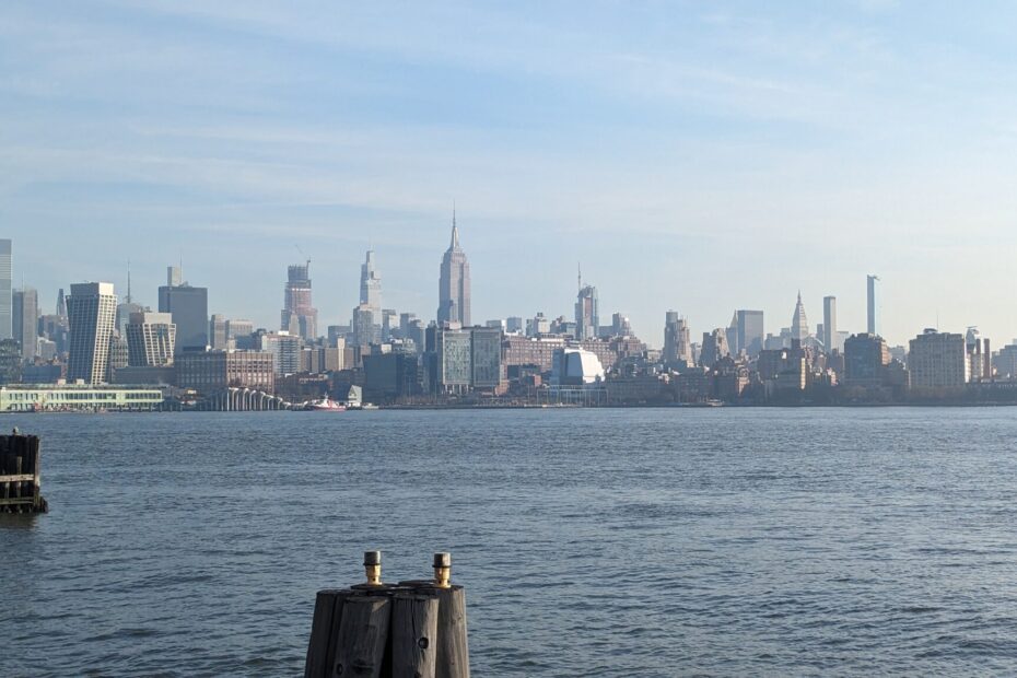 It is the photo presenting a clear view across the Hudson River towards Midtown Manhattan with the Empire State Building prominently visible. This iconic skyline is characterized by its diverse range of architectural styles and the towering presence of skyscrapers, forming a distinctive and instantly recognizable urban landscape. The river in the foreground adds to the scenic beauty of the view, with the water acting as a natural mirror to the city's grandeur.