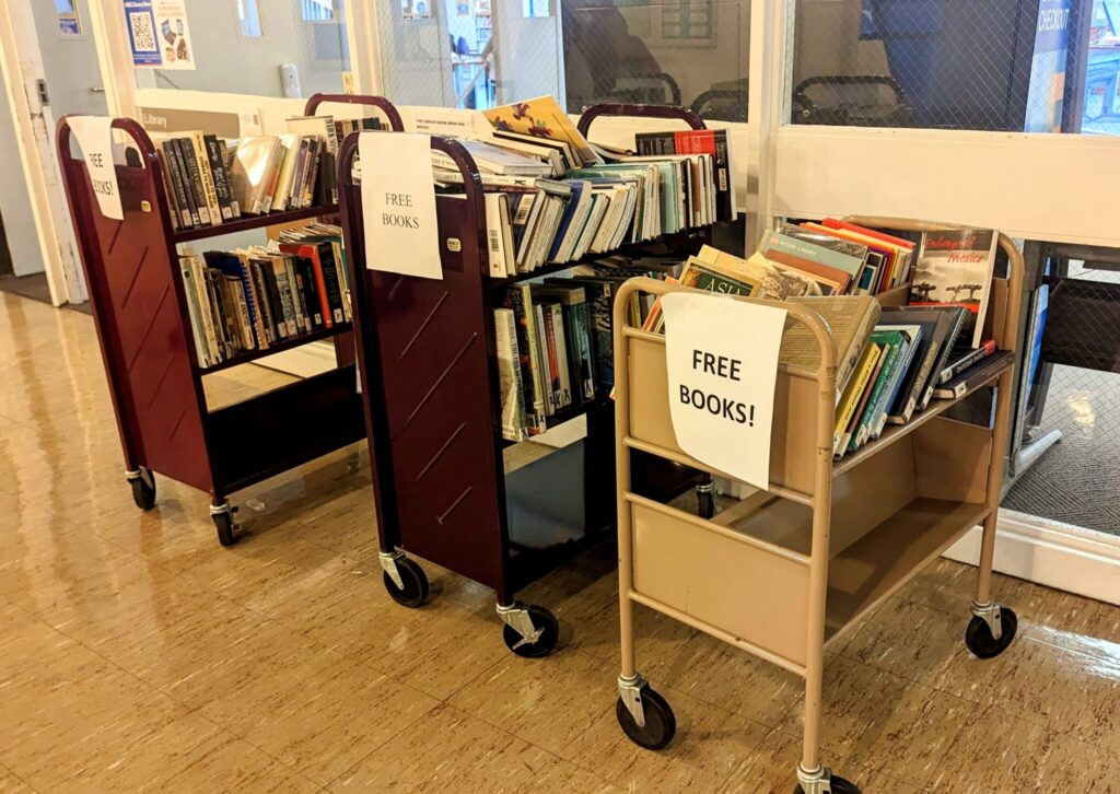 Three metal carts full of books. On each cart is taped a sign which reads, "Free Books!"